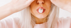 Closeup of the mouth of a blonde young woman blowing a pink gum bubble while wearing a pink t-shirt